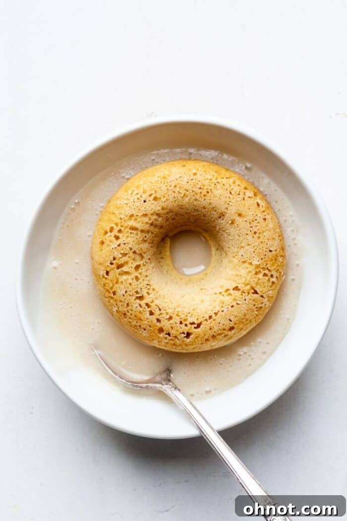 A freshly baked gluten-free donut being dipped into the creamy vanilla glaze.