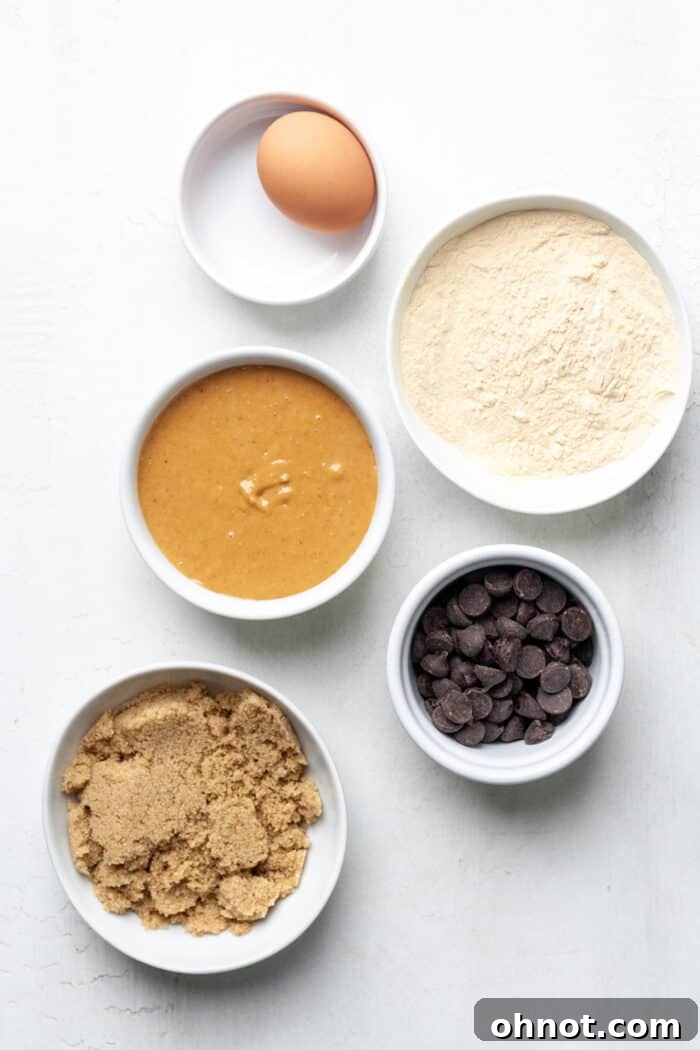 Individual ingredients for protein chocolate chip cookies displayed in small white bowls, including peanut butter, egg, brown sugar, protein powder, and dark chocolate chips.