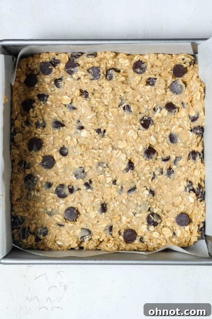 Raw oatmeal chocolate chip bar dough spread evenly in a parchment-lined baking pan.