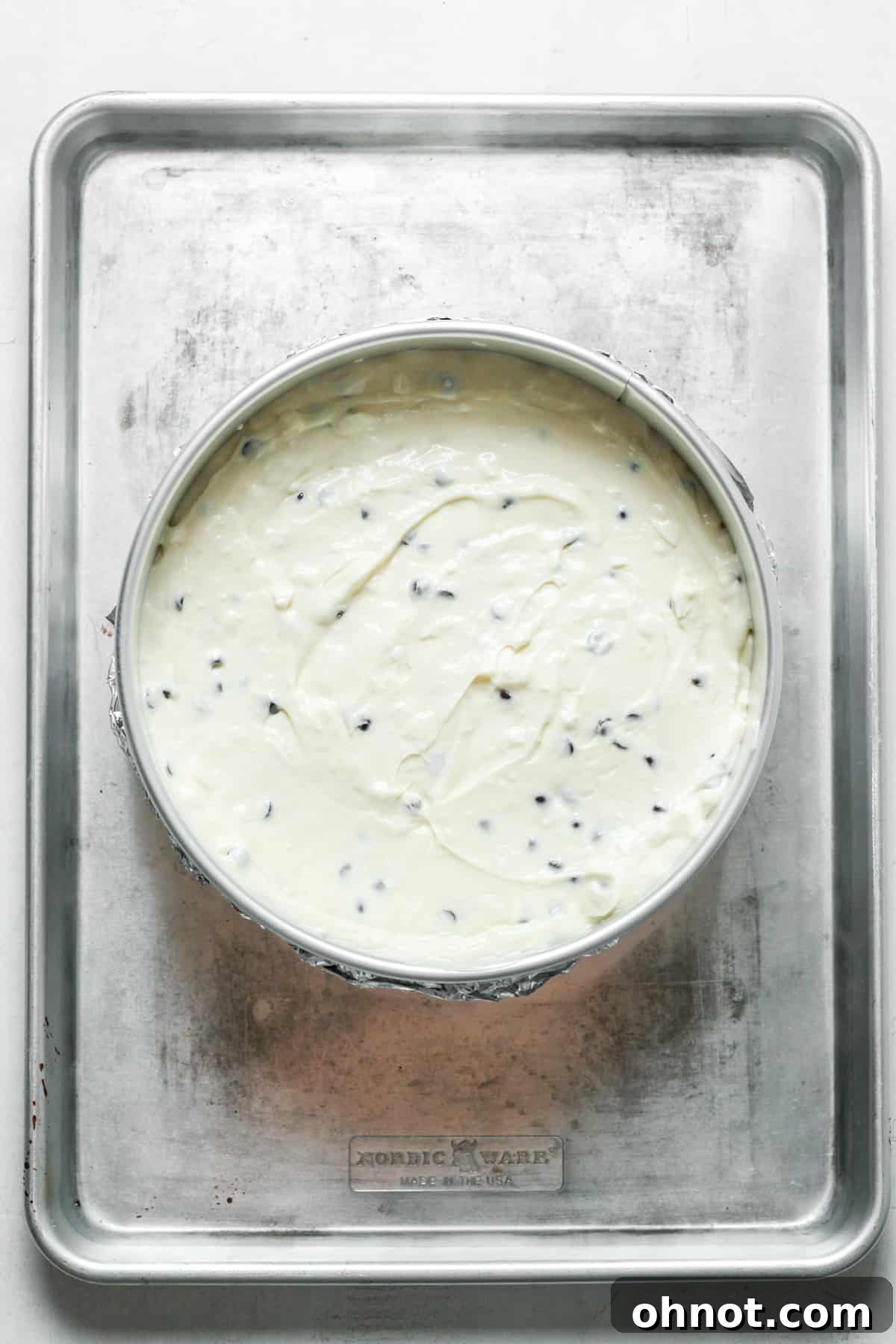 A springform pan with cheesecake filling, wrapped in foil, placed in a roasting pan for a water bath.