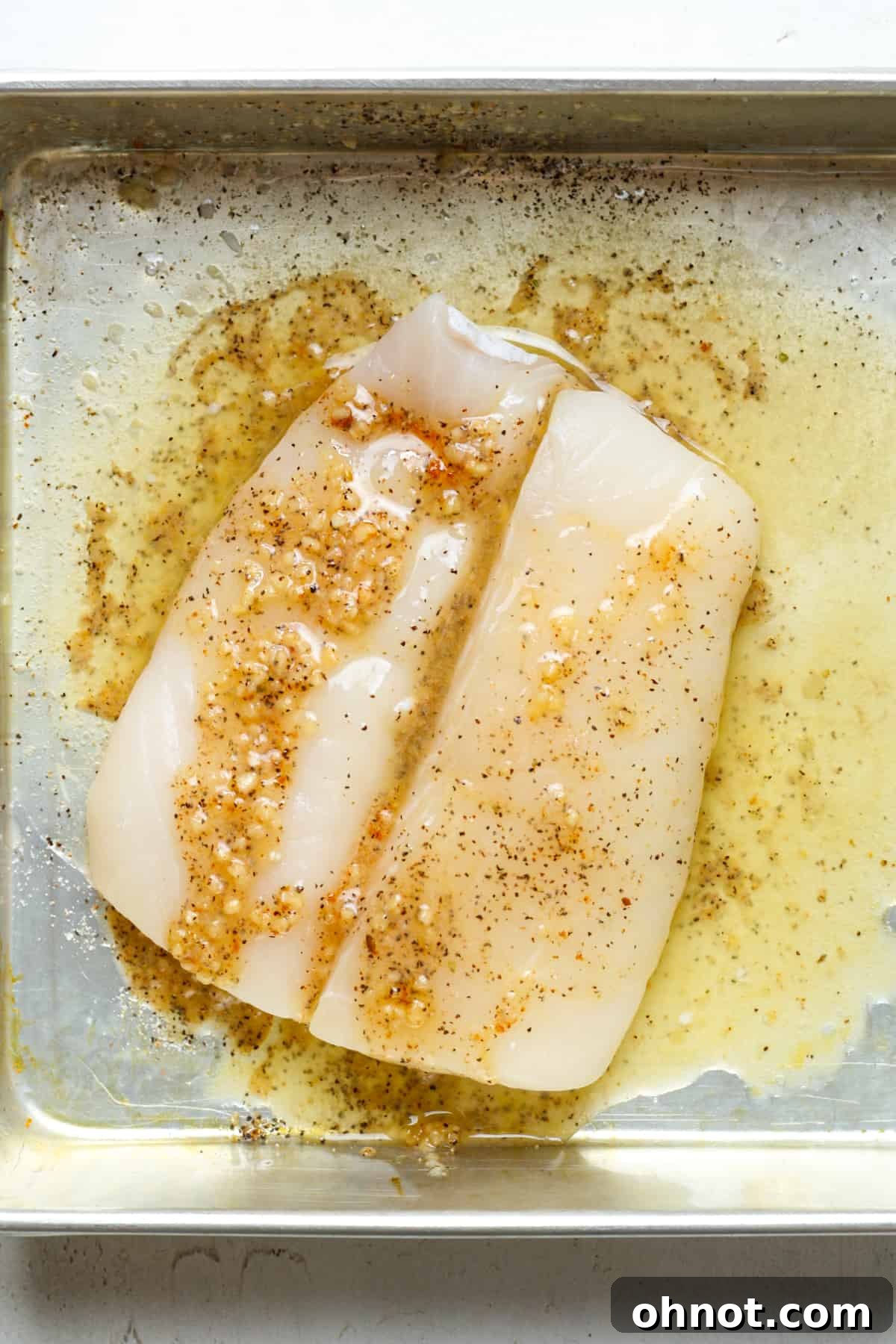 A halibut fillet in a baking dish, generously being drizzled with the prepared garlic butter sauce.