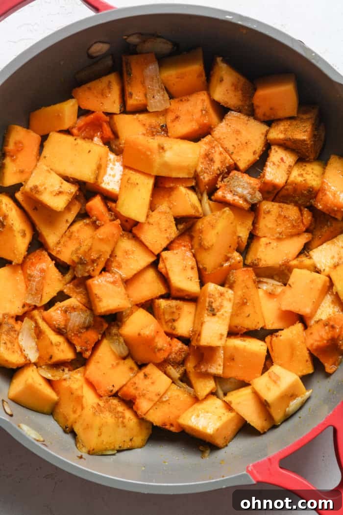 Cubed butternut squash and spices being stirred into sautéed onions in a skillet.