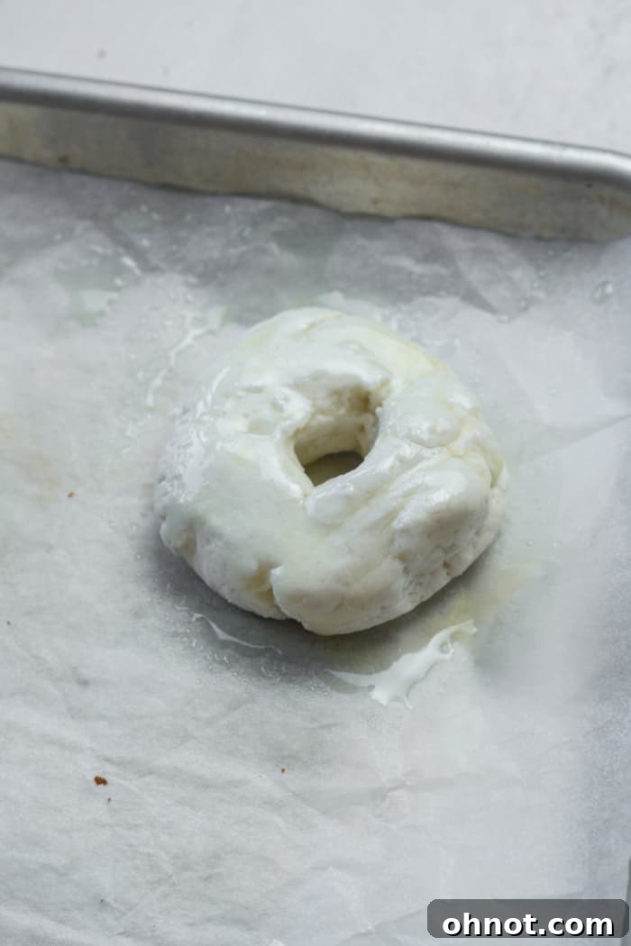 Nonstick spray being applied generously to raw bagels on a baking sheet.