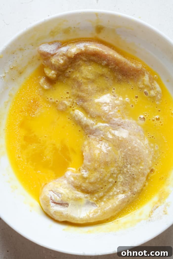 Chicken breast dipped in whisked egg, showcasing the second stage of the breading process.