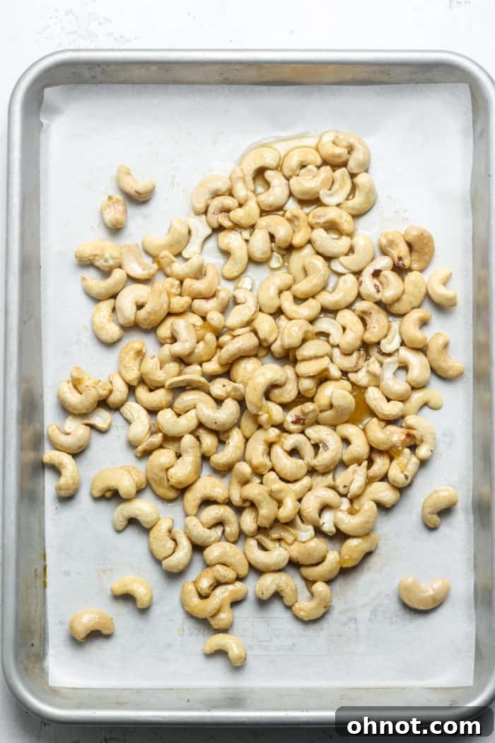 Honey-coated cashews spread in a single, even layer on a parchment-lined baking sheet, ready for roasting.