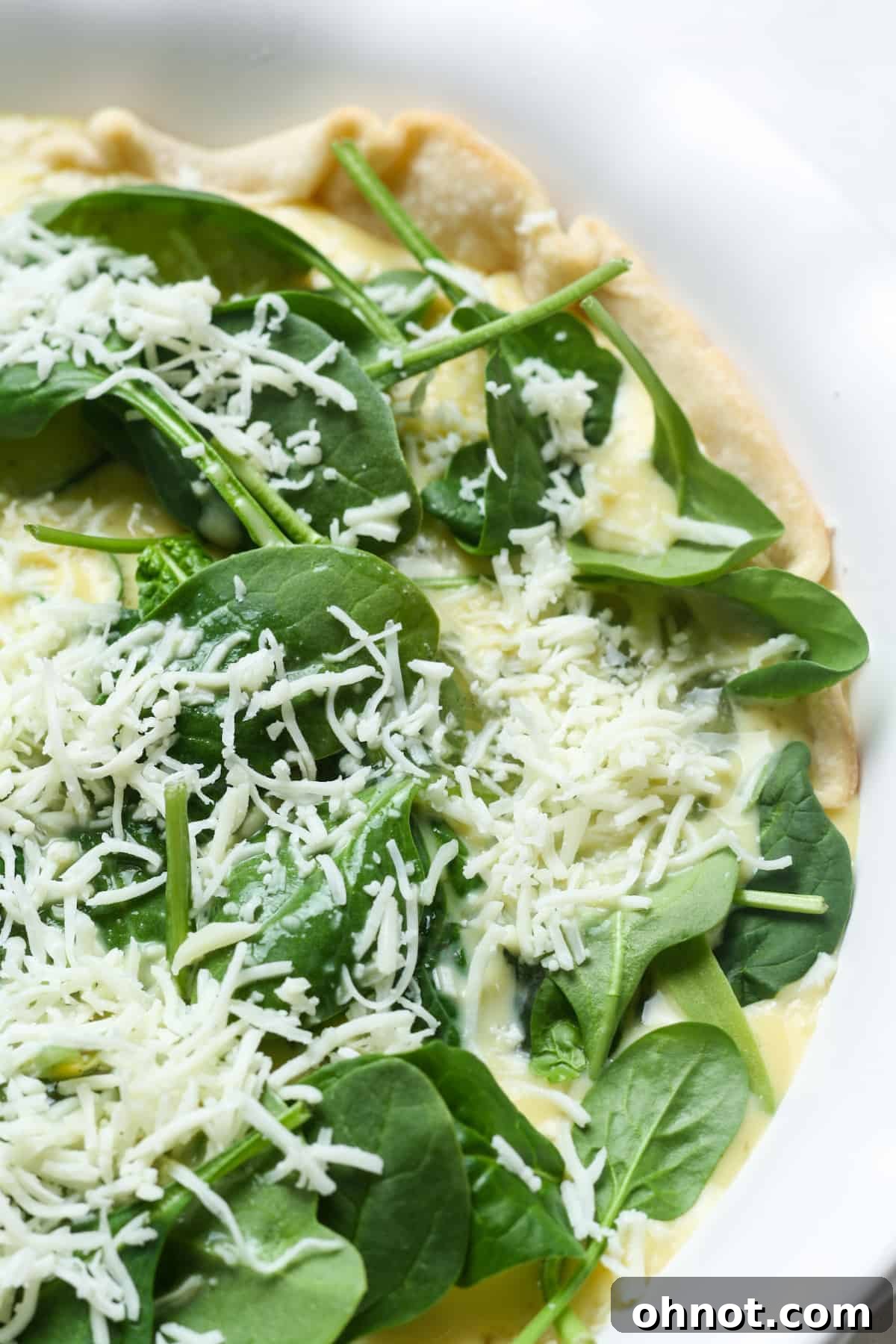 A pie crust filled with the egg custard, spinach, and cheese, topped with more cheese, ready for baking.