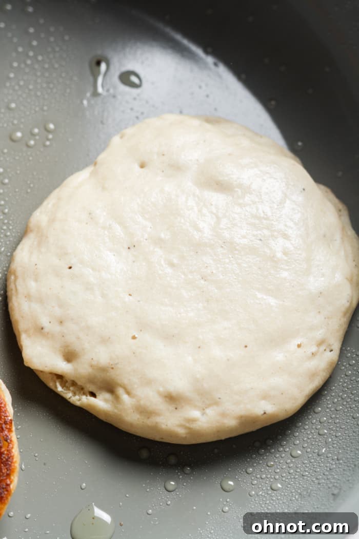 A perfectly circular pancake batter cooking on a hot buttered skillet, showing small bubbles forming on the surface.