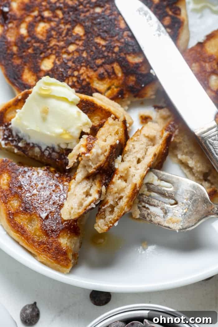 A breakfast plate featuring a stack of sourdough pancakes, a bite taken out of one, alongside fresh fruit like berries and sliced banana, and a drizzle of maple syrup.