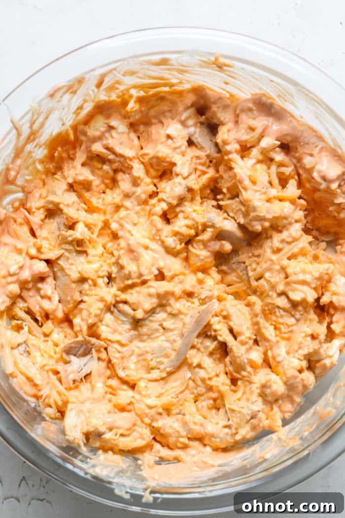 A bowl filled with spicy chicken, creamy sauce, and shredded mozzarella cheese, mixed thoroughly.