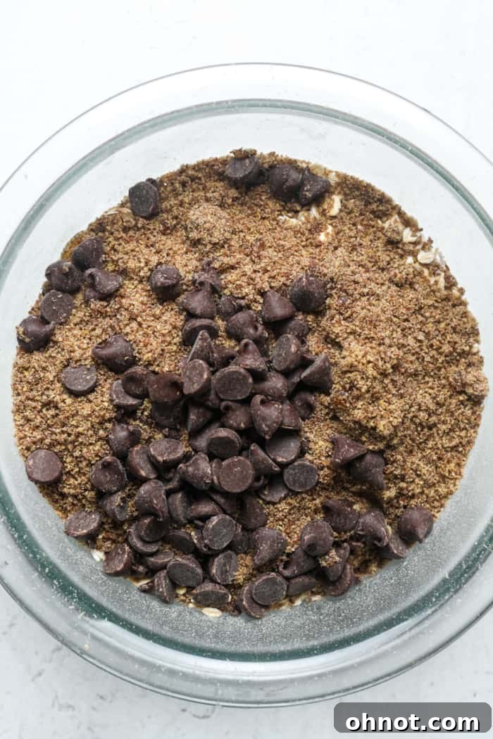 Ground flaxseed and chocolate chips being added to a mixing bowl with oats and peanut butter.