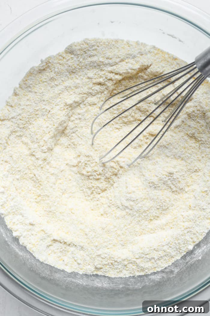 A close-up view of a mixing bowl containing the dry ingredients for cornbread, showing the white gluten-free flour and yellow cornmeal blended together.