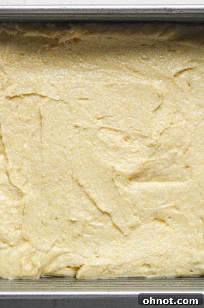Thick, yellow cornbread batter neatly spread into a square baking pan, ready to be placed in the oven for baking.