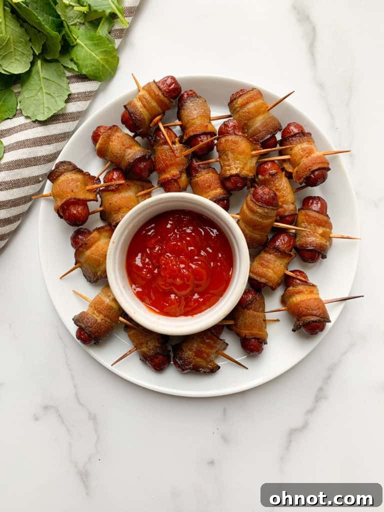 A simple white plate features a neat arrangement of mini hot dogs, expertly wrapped in crispy bacon, with a small dollop of ketchup on the side.