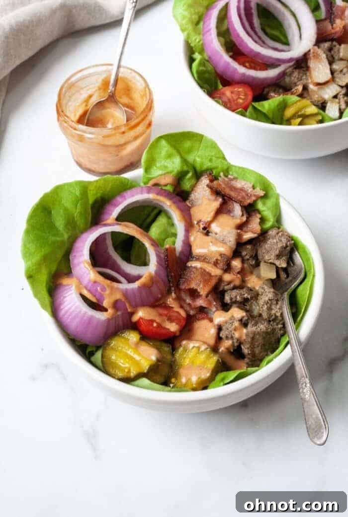 A clean white bowl is brimming with a vibrant mix of perfectly seasoned ground beef, crisp lettuce, thinly sliced onions, juicy tomatoes, and crunchy pickles, forming a wholesome burger bowl.