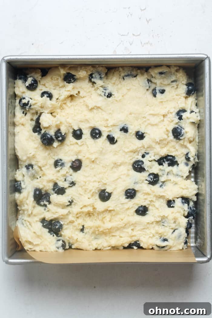 The blueberry coffee cake batter evenly spread in a parchment-lined square baking pan, ready for the streusel topping.