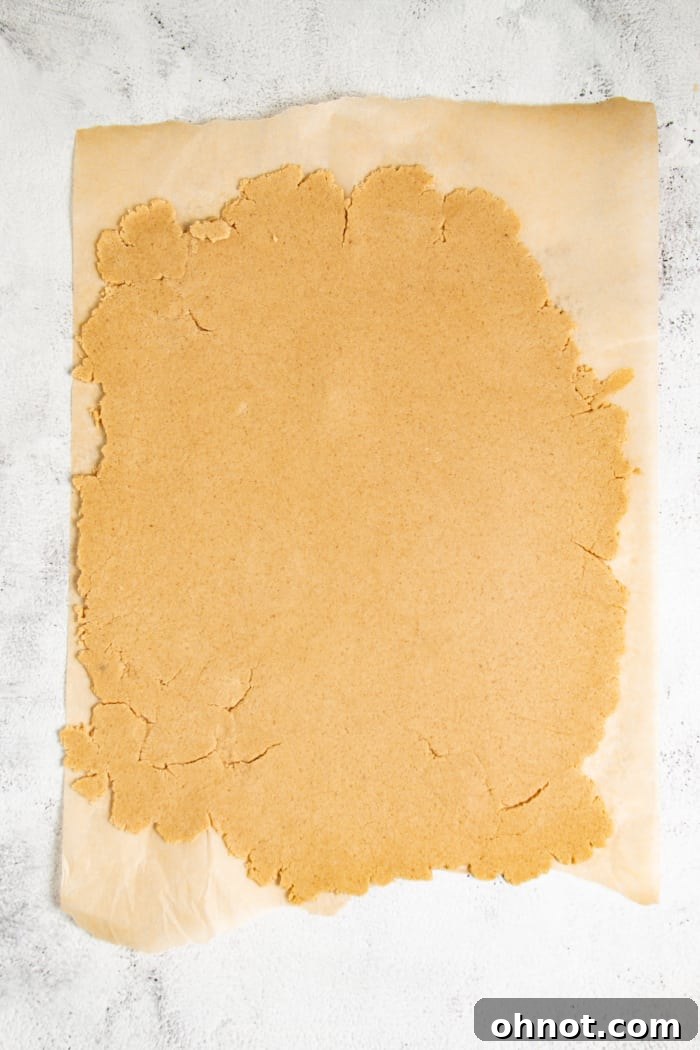 Graham cracker dough rolled out thinly between two sheets of parchment paper, with a rolling pin visible, demonstrating the preparation stage before cutting.