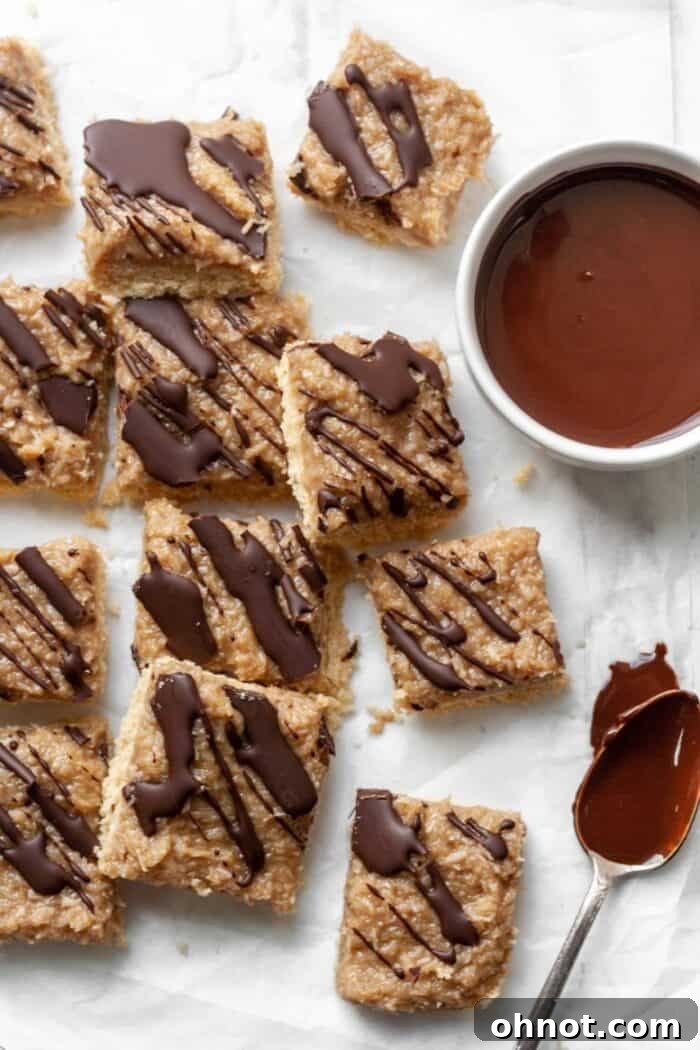 Close-up of three delicious Paleo Samoa bars stacked neatly on white parchment paper, with a bowl of melted chocolate in the soft-focus background, ready for drizzling.