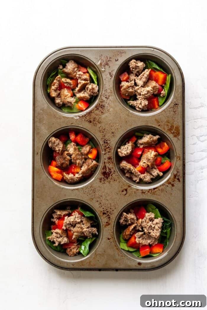 A muffin tray filled with layers of spinach, red peppers, and cooked sausage, ready for the egg mixture.