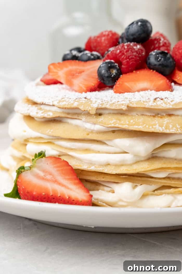 A stunning, multi-layered Crepe Cake garnished with fresh berries and a light dusting of powdered sugar, presented on a minimalist white cake stand.