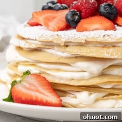 A stunning, multi-layered Crepe Cake garnished with fresh berries and a light dusting of powdered sugar, presented on a minimalist white cake stand.