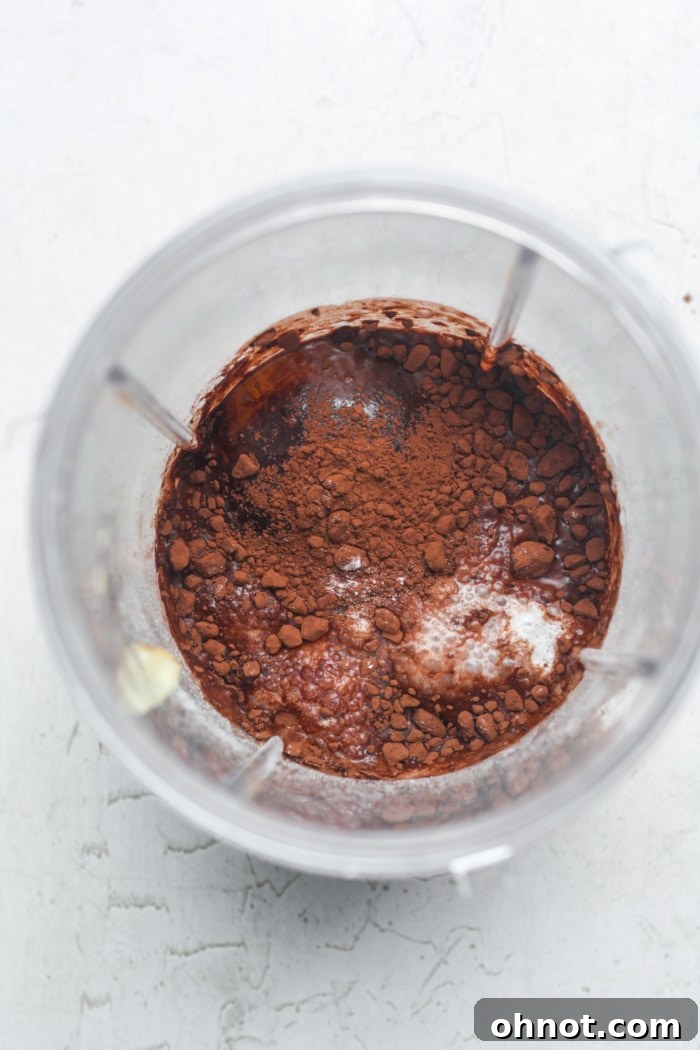 A blender jar with cocoa powder being added, surrounded by other ingredients for baked oats.