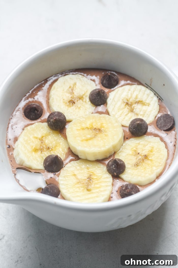 An oven-safe bowl filled with chocolate oat batter, garnished generously with fresh banana slices and dark chocolate chips, ready for baking.