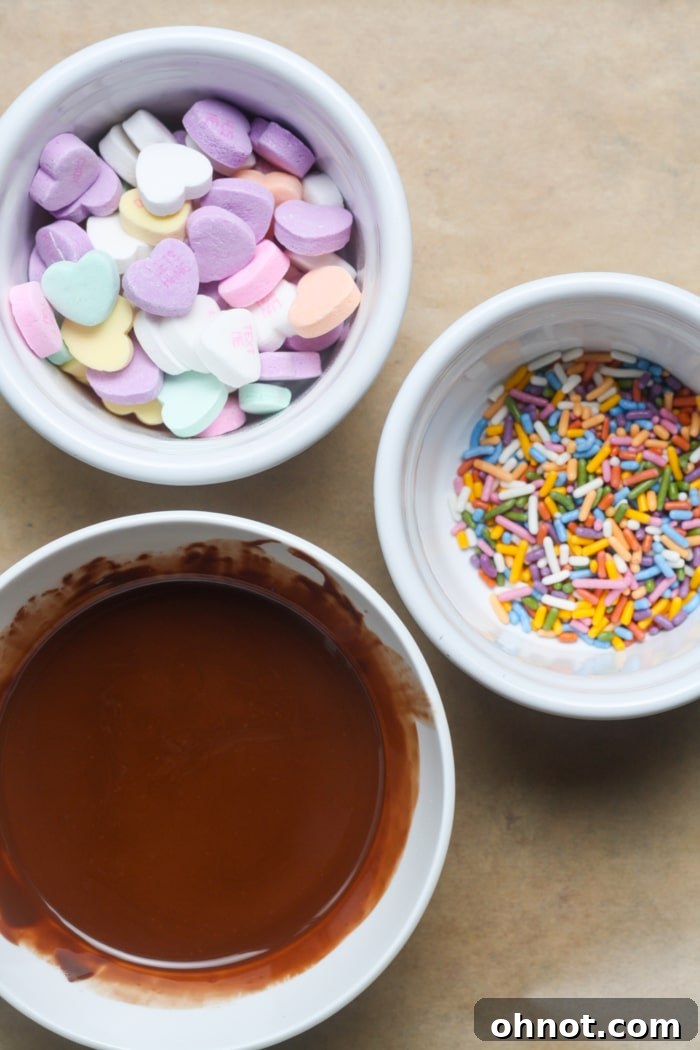 A collection of ingredients for Valentine's bark, including chocolate chips, coconut oil, conversation hearts, and sprinkles, laid out on a surface.