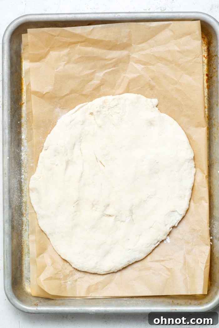 A flat, round raw pizza crust stretched out on parchment paper on a baking pan, ready for its initial bake.