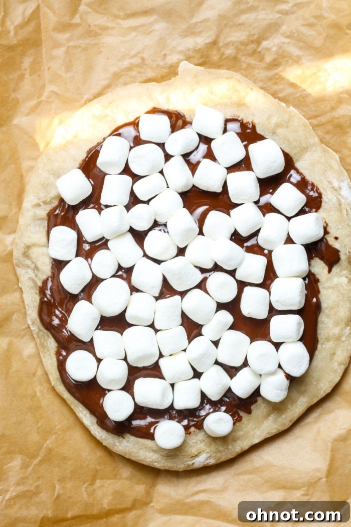 A pre-baked pizza crust covered with a layer of melted chocolate, topped generously with mini marshmallows, awaiting its final bake.