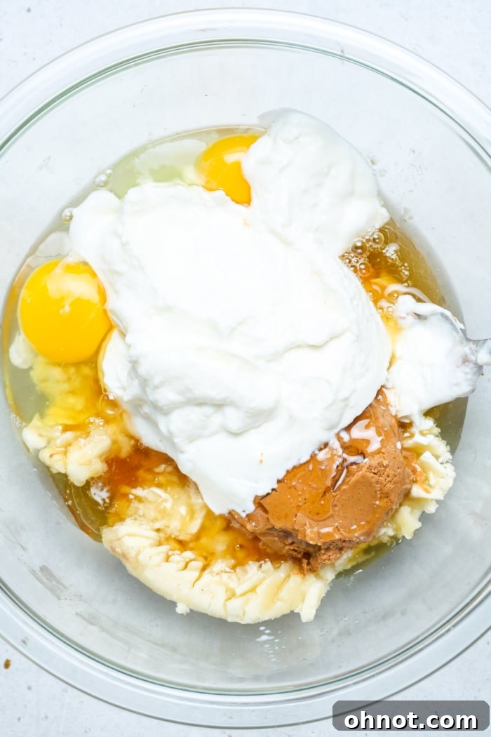 A large mixing bowl containing mashed bananas, eggs, Greek yogurt, and peanut butter, ready to be combined.