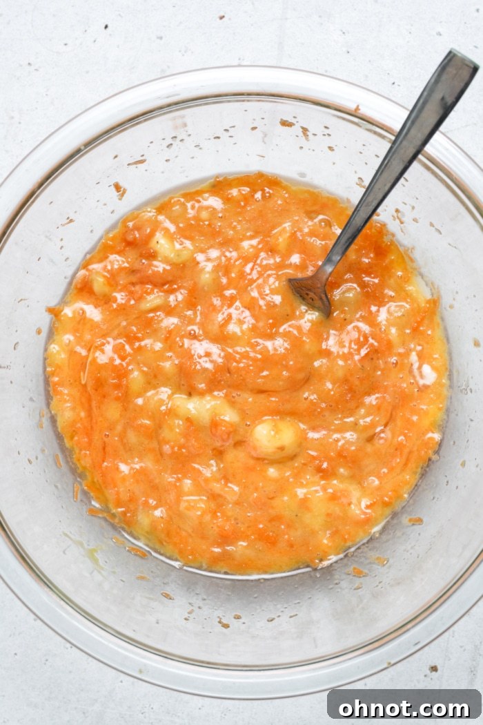 Vibrant orange batter in a large mixing bowl, showcasing the mashed bananas, shredded carrots, eggs, and coconut oil mixed together.