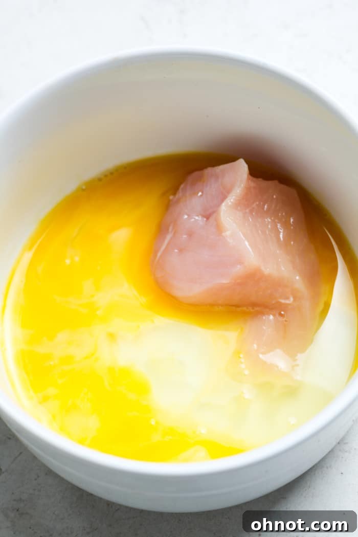 A piece of chicken breast being dipped into a shallow bowl of whisked egg wash.