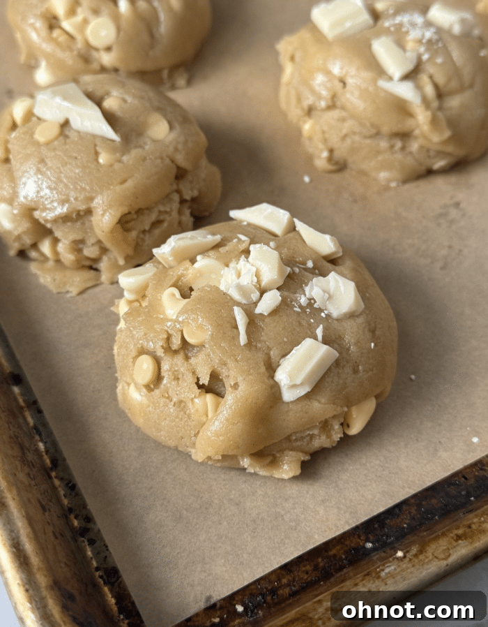 Unbaked cookie dough balls with white chocolate chunks on a parchment-lined baking sheet, ready for chilling.