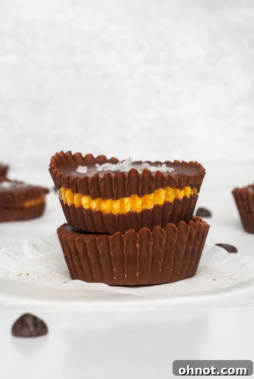 Two pumpkin chocolate cups on a white plate, showcasing the chocolate base, pumpkin filling, and chocolate topping with a sprinkle of sea salt. The image highlights the beautiful layers and inviting texture of the no-bake treats.