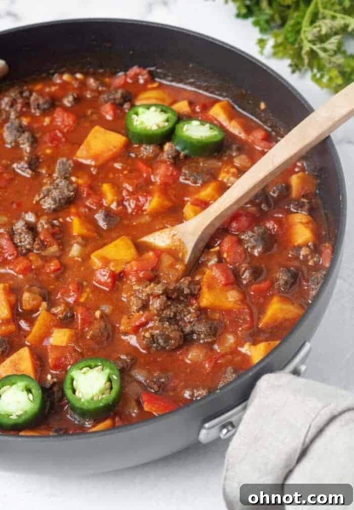 A large black skillet filled with simmering Whole30 Sweet Potato Chili, stirred with a wooden spoon.