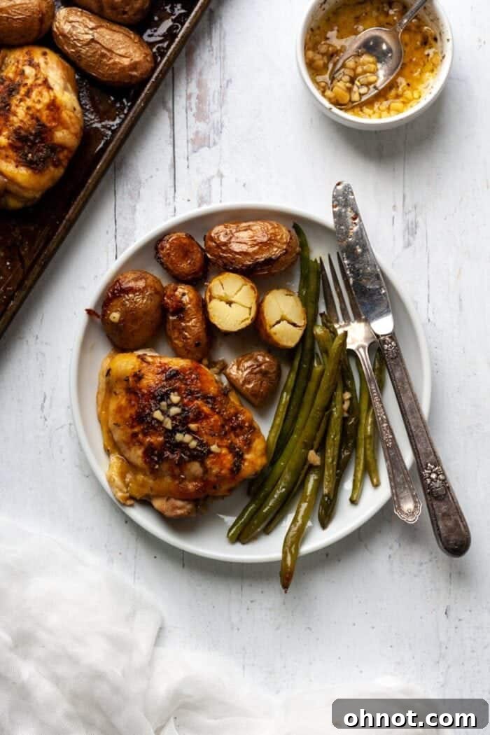 A top-down view of a baking pan filled with golden-brown chicken thighs, roasted fingerling potatoes, and vibrant green beans, seasoned with herbs.