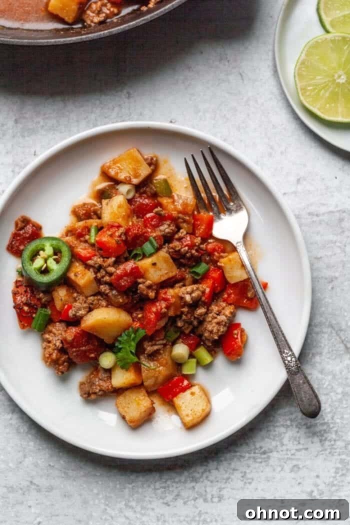 A beautifully plated Paleo Taco Skillet, showcasing vibrant ingredients like ground beef, potatoes, and fresh garnishes, set on a white plate beside a silver fork and lime wedges.