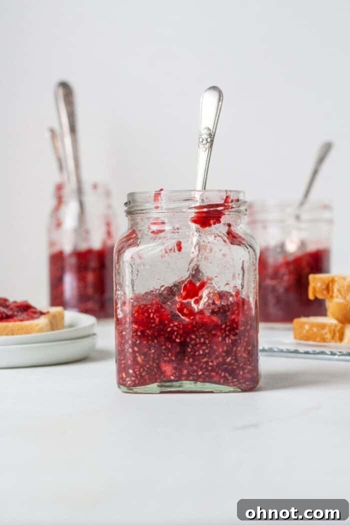 A beautifully clear glass jar filled with homemade, vibrant red sugar-free raspberry chia jelly, showcasing the tiny chia seeds and fresh raspberries. The jar sits on a rustic wooden surface, hinting at natural goodness.