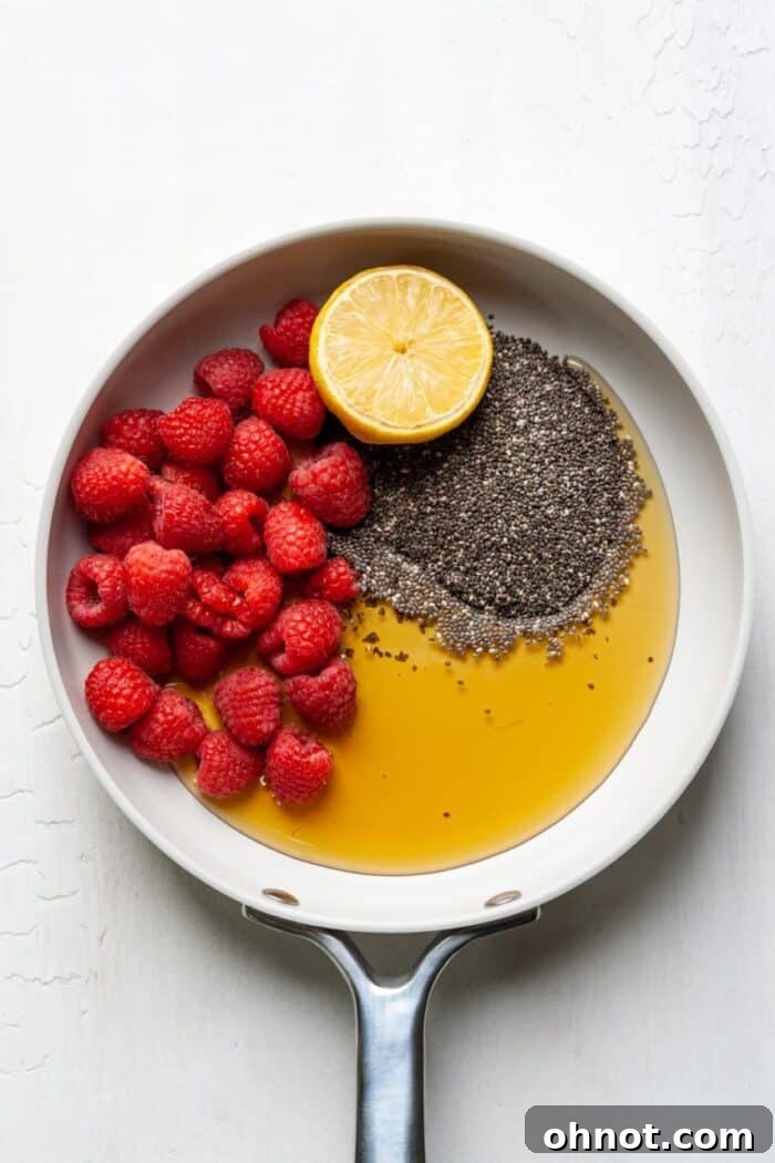 A bright white non-stick skillet on a stovetop, filled with fresh red raspberries, tiny black chia seeds, and a liquid mixture of maple syrup and lemon juice, just beginning to warm up.