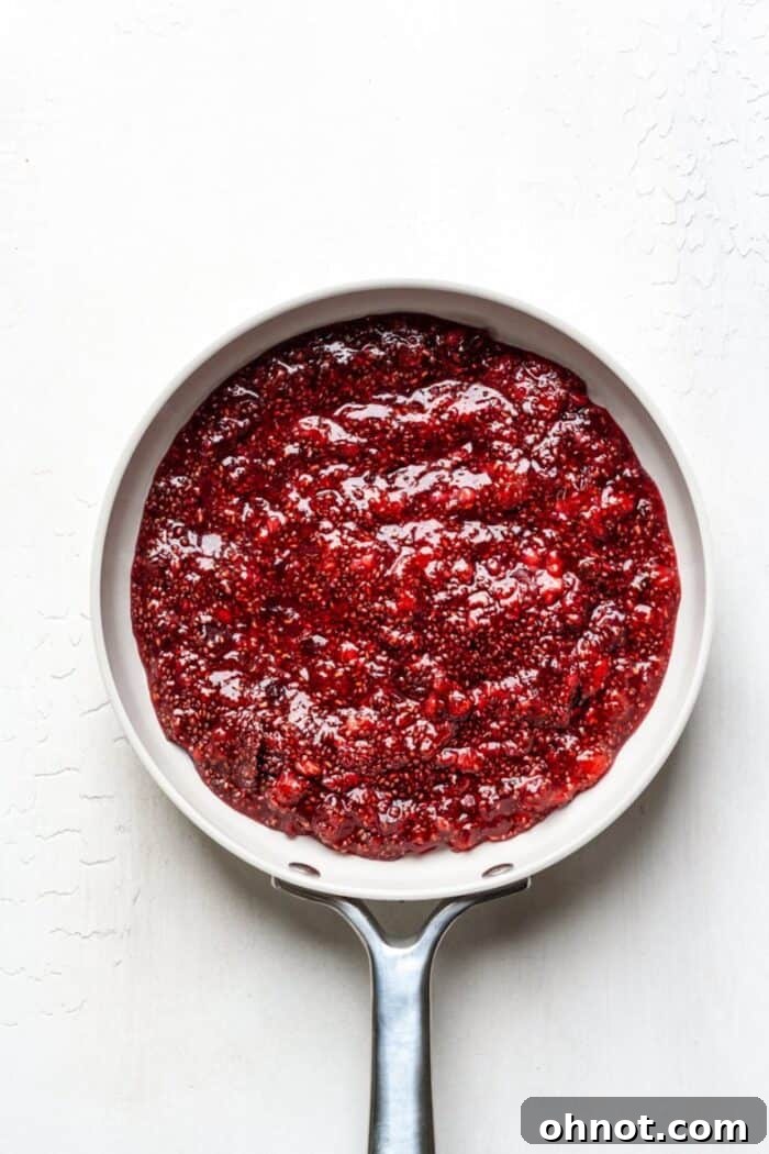 A close-up shot of the white skillet on a stovetop, now filled with a vibrant red, thickened vegan raspberry chia jelly, showing the bubbling texture and the plump chia seeds integrated into the fruit spread.