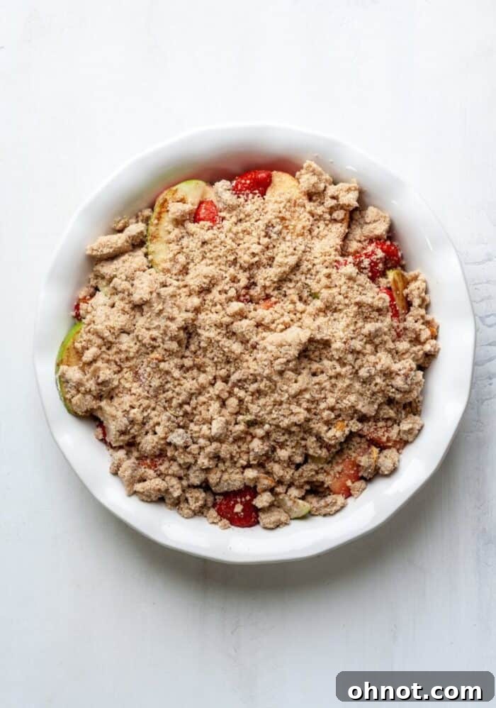 An unbaked apple strawberry crumble in a white pie dish, topped with the crumble mixture and ready for the oven.