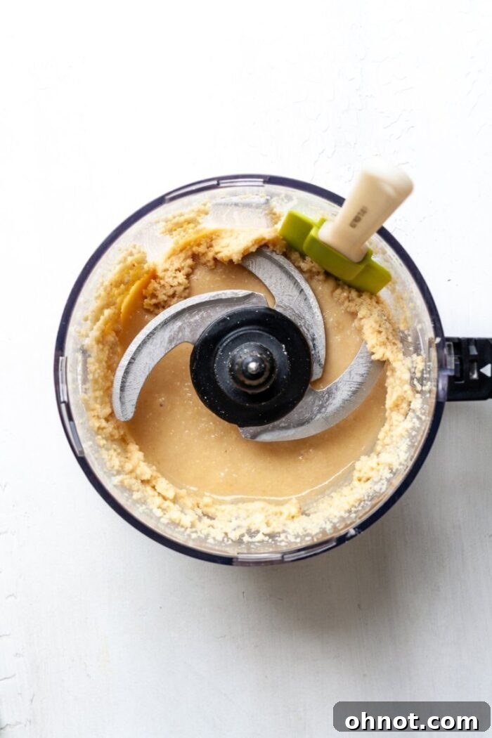 A food processor containing partially blended macadamia nuts, with a spatula scraping down the sides.